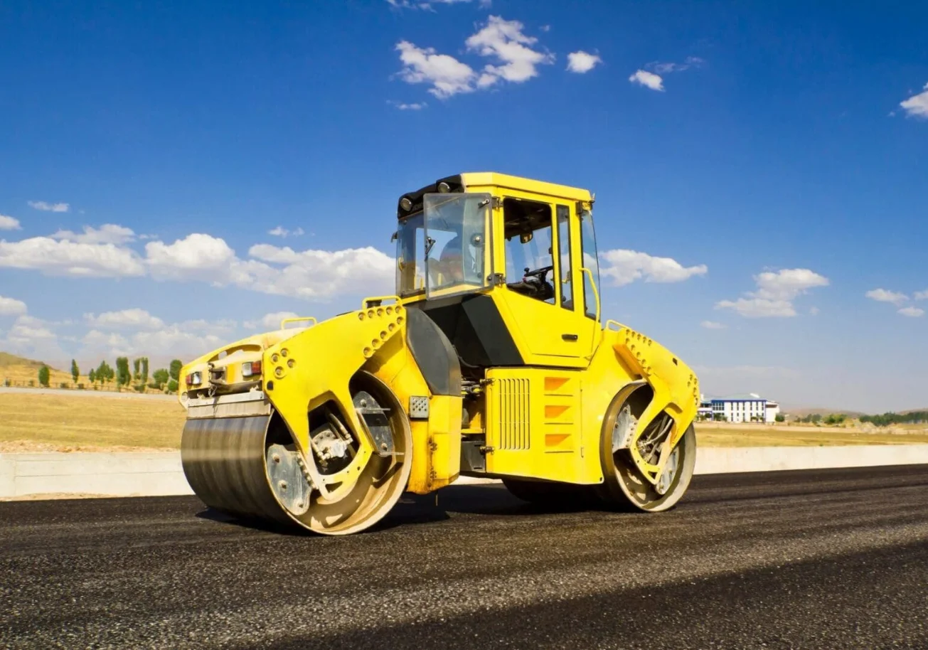 Freshly paved asphalt driveway on a residential property in Sonoma County, California, with a smooth dark surface and clean edges bordered by dry grass and oak trees. A paving crew operates a compaction roller in the background during the finishing stage.